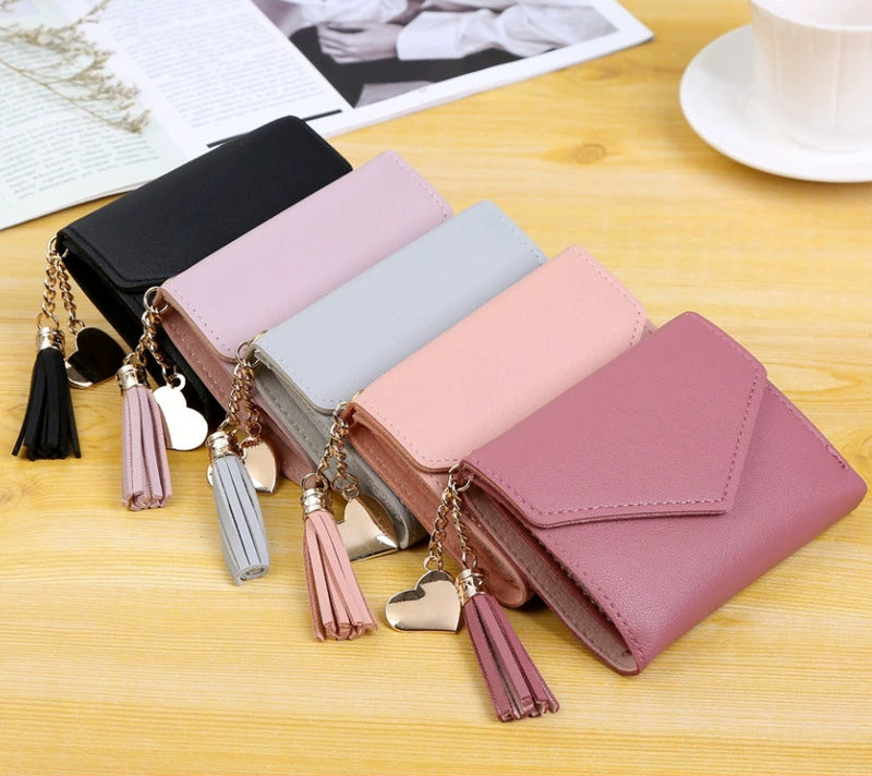 Five small zip-around wallets in black, pale pink, light blue, blush pink, and dusty rose are arranged on a light wood surface. Each wallet has a tassel charm with a heart-shaped metal pendant.