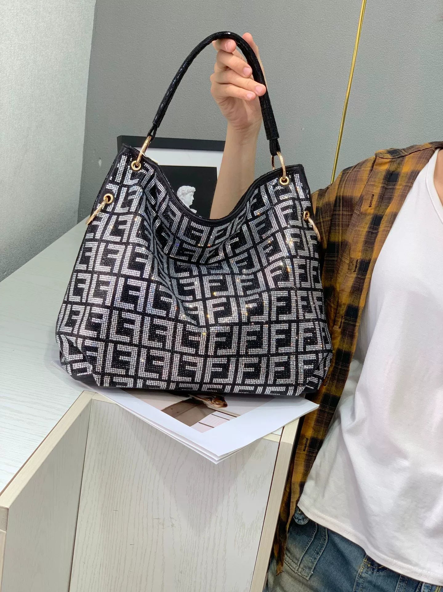 A person holds up a Fendi shoulder bag with a black and white FF monogram pattern, featuring a black leather top handle and gold-tone hardware, set against an indoor background.
