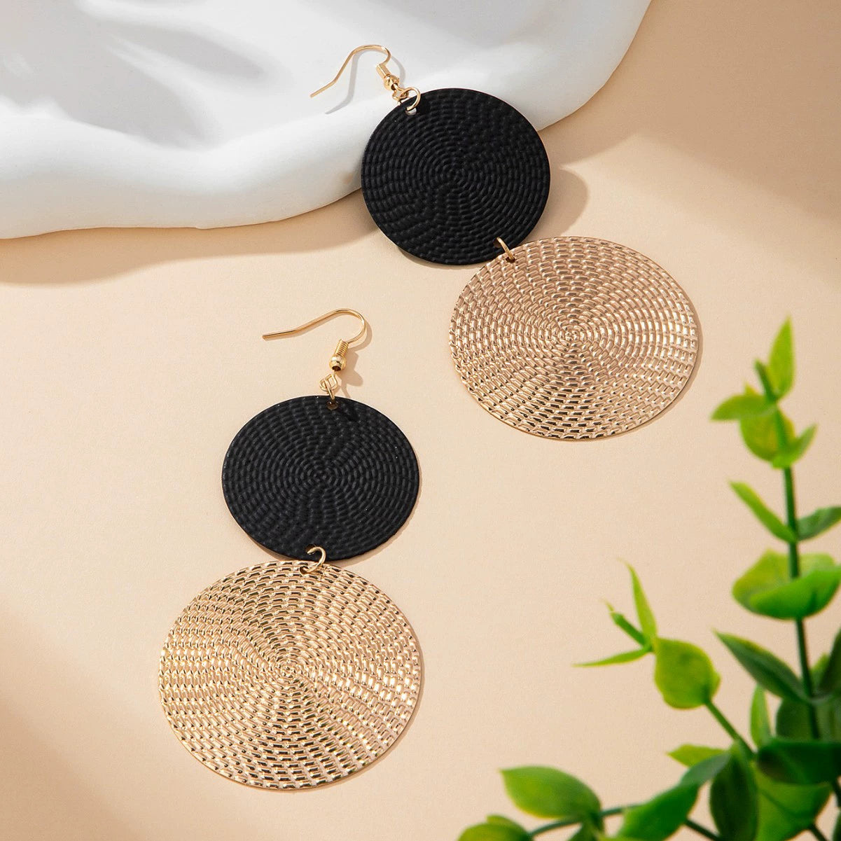 Pair of black and gold circular earrings on a beige surface with a plant in the corner.